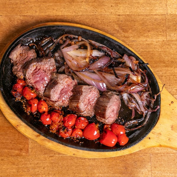 Sizzling steak pieces, roasted red onions, and cherry tomatoes on a black cast iron platter.