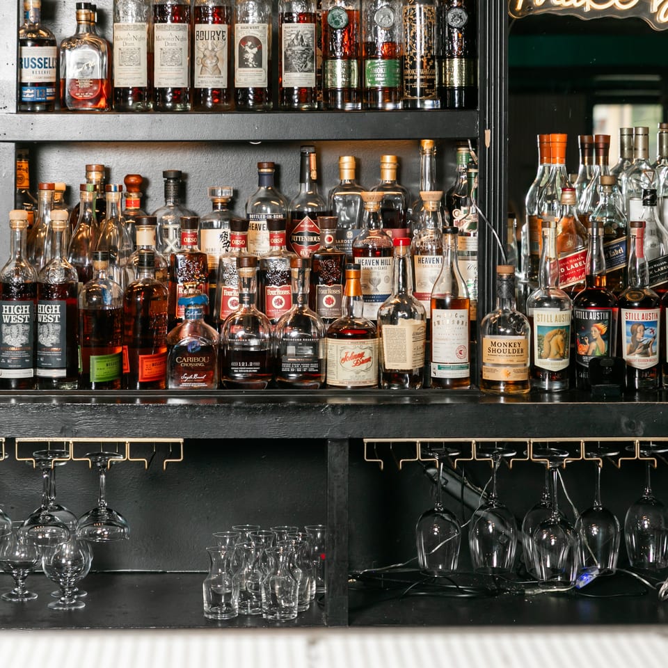 A well-stocked bar shelf displays numerous bottles of liquor, with hanging glasses below.