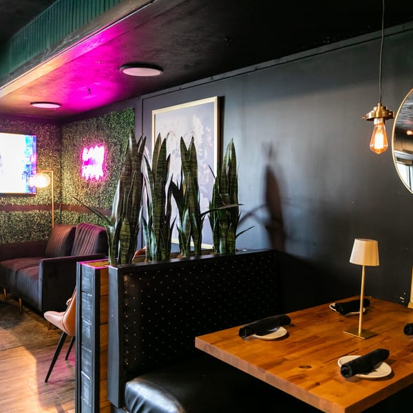 A restaurant interior with a booth, wooden table, modern lighting, and a lounge area with a TV.