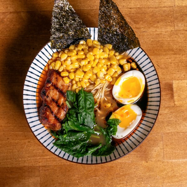 Ramen bowl with grilled pork, corn, spinach, soft-boiled eggs, and nori on a wooden table.