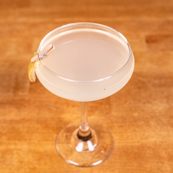 Light-colored cocktail in a coupe glass, garnished with a lemon peel held by a tiny clothespin.