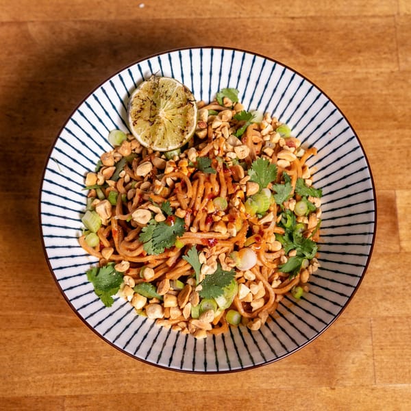 A bowl of noodles with peanuts, cilantro, green onions, and a grilled lime wedge.
