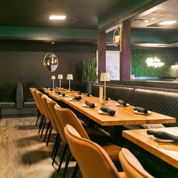 Restaurant interior with long wooden tables, brown chairs, black booths, and dark green walls.