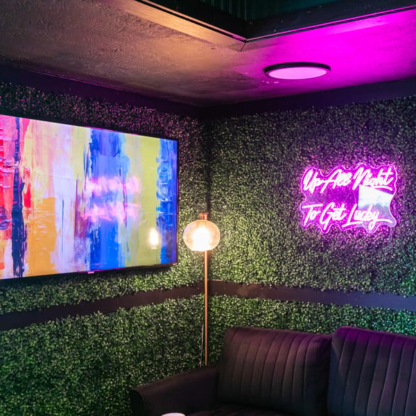 A lounge area with a green hedge wall, a colorful abstract TV, and a neon sign reading "Up All Night To Get Lucky".