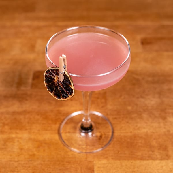 Pink cocktail in a coupe glass, garnished with a dried citrus slice on a wooden table.