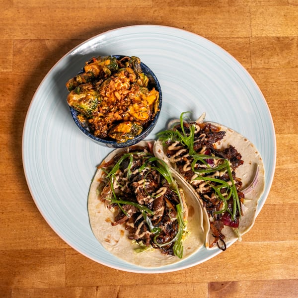Two tacos with meat, onions, and green garnish, alongside a bowl of roasted Brussels sprouts.