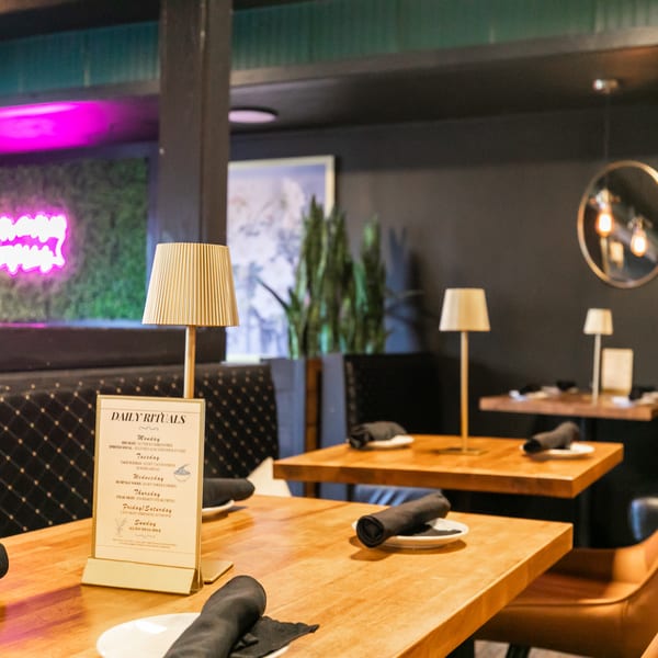 A restaurant interior with wooden tables, black chairs, a neon sign, and table lamps.